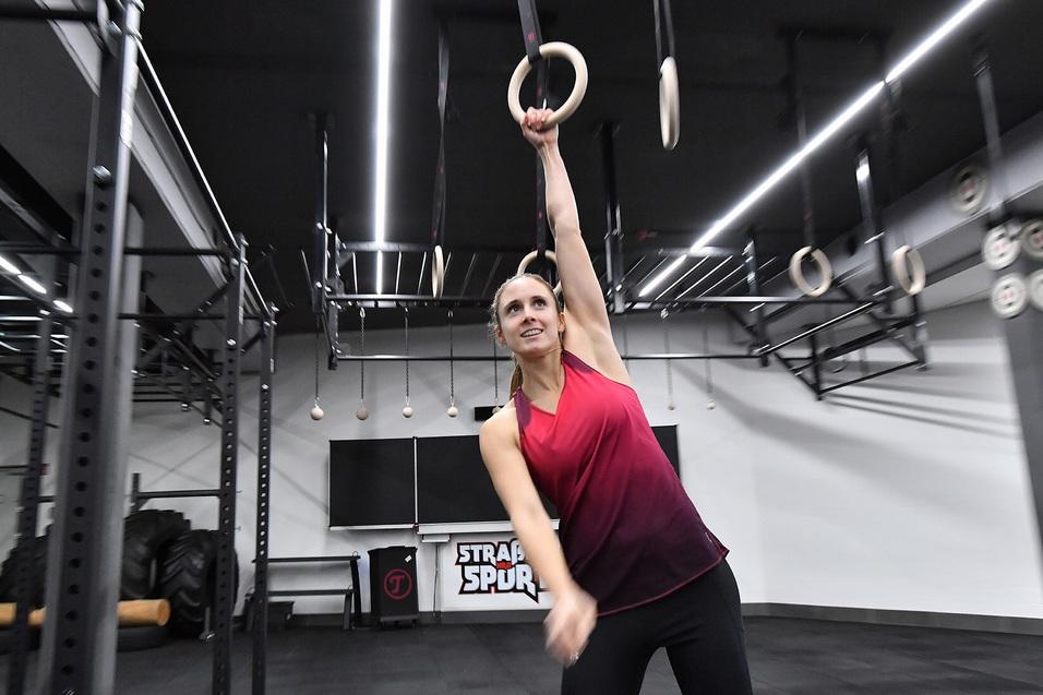 Frau in sportlicher Kleidung greift mit einer Hand nach Turnringen in einem modernen Fitnessstudio.