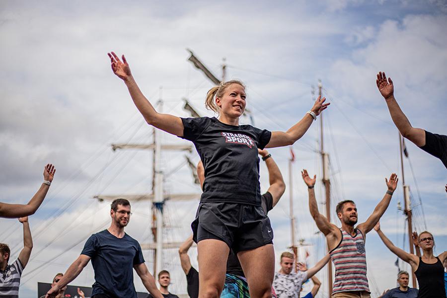 Eine Gruppe von Menschen macht gemeinsam Fitnessübungen vor historischen Segelschiffen unter einem bewölkten Himmel.